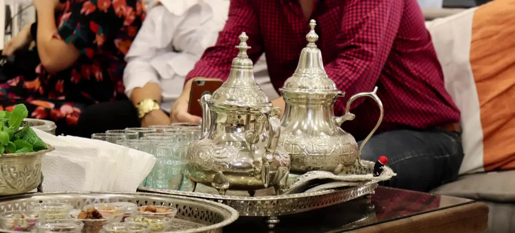مجموعة من الأشخاص يجلسون حول طاولة يستمتعون بالشاي المغربي يقدمون من أباريق الشاي الفضية.